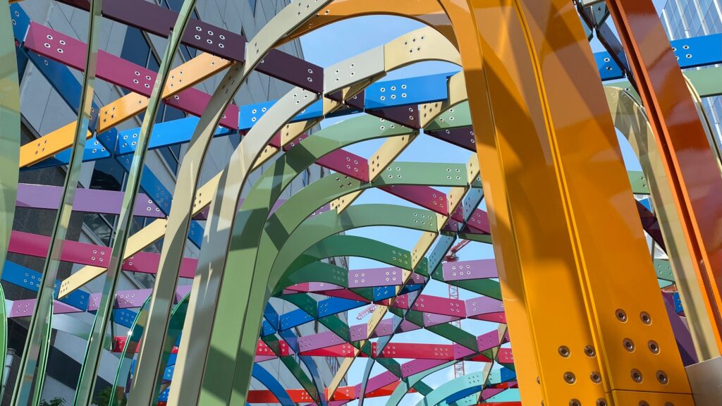View of a metal sculpture from below with buildings and sky visible through it. The sculpture pieces are attached overhead like an arbor, and are multi-colored.