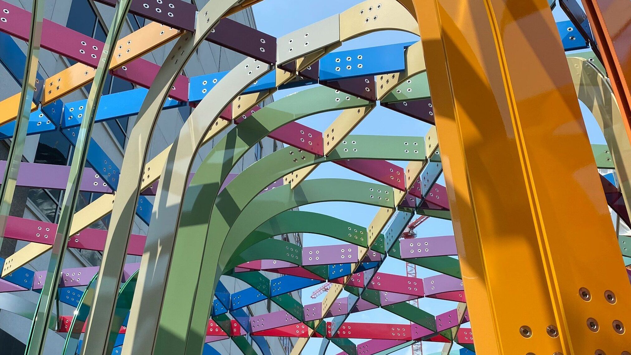 View of a metal sculpture from below with buildings and sky visible through it. The sculpture pieces are attached overhead like an arbor, and are multi-colored.