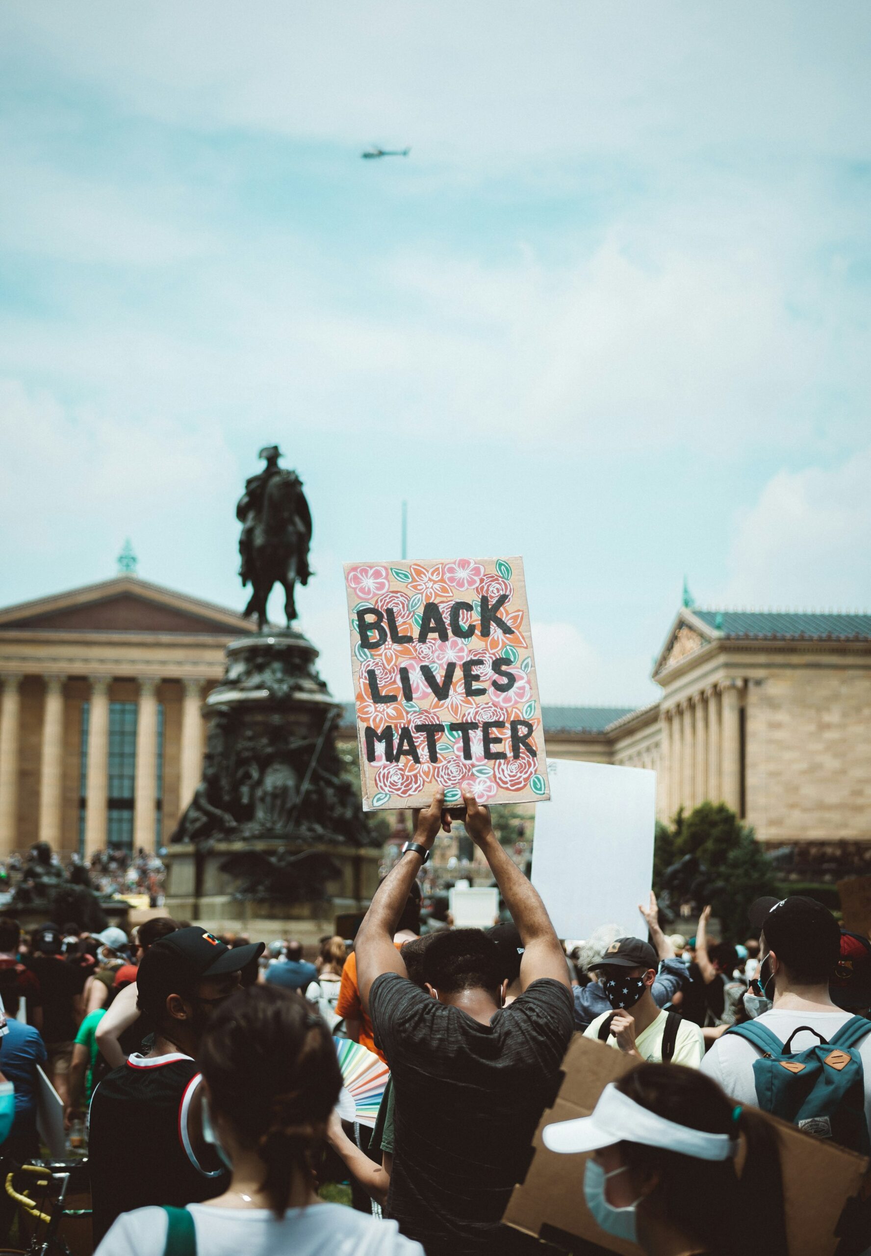 A group of demonstrators are gathered in front of the Washington Monument at the Philadelphia Museum of Art. Most face away from the photographer. One man is looking down and holding a sign that reads "BLACK LIVES MATTER" above his head with both hands. The background of the sign is illustrated with pink, orange, and red flowers, some roses, and green leaves. In the sky, a helicopter is visible but blurry.