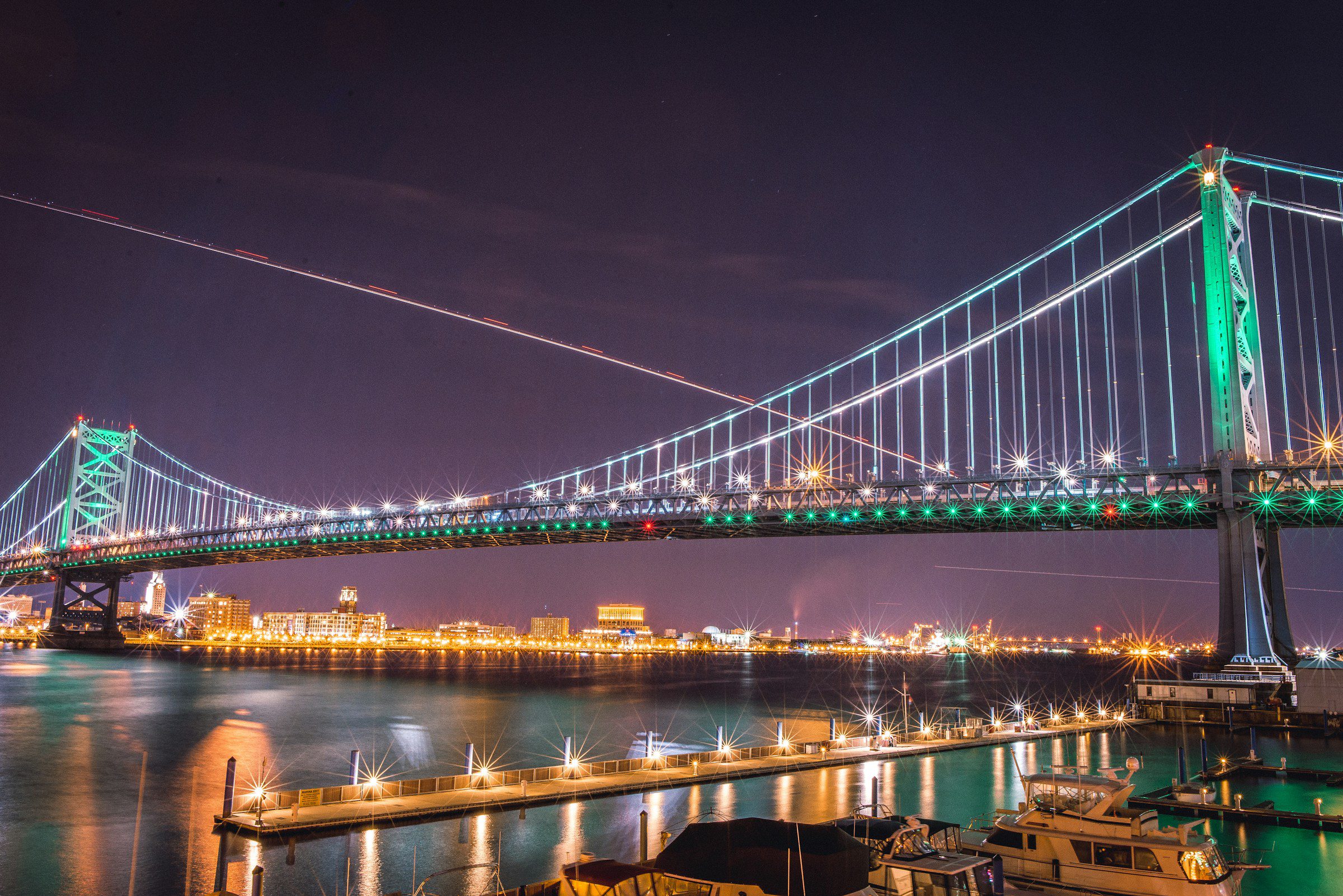 Long exposure of Ben Franklin Bridge in Philadelphia at night with lights. The sky is purple and has visible trails from aircraft.