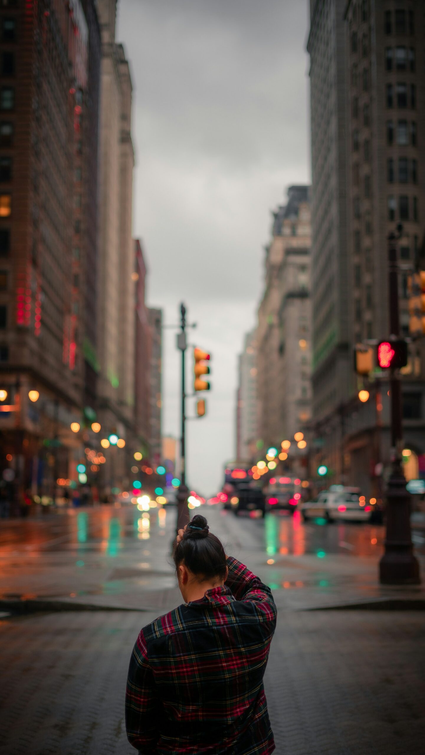 A woman in a plaid shirt facing away from the camera looks down at the sidewalk near a city street. Her right hand is on her head, possibly adjusting her hair, which is wrapped in a bun. The street is busy with cars, and the weather is cloudy and foggy. The lights reflect in the wet street. Tall buildings flank both sides of the street, but the sky is visible in the center, cloudy and gray.