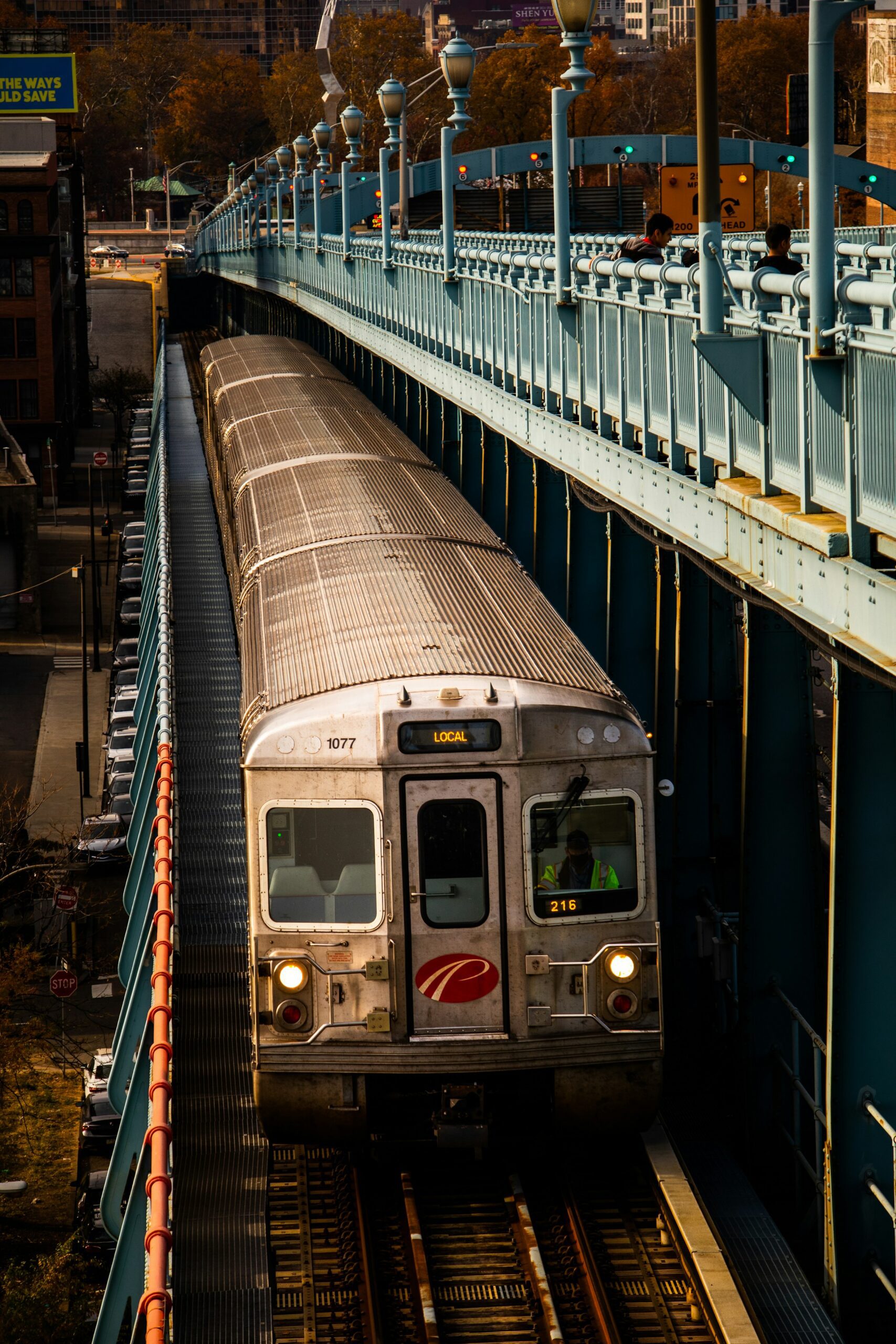 A silver PATCO commuter train is traveling toward the viewer on a raised track. The train is labeled "LOCAL" and the conductor is visible through the front glass. The conductor wears an orange and yellow safety vest. Above the train, a bridge for pedestrians and cars is visible. It has blue railings and is lined with streetlights. Two men are standing on the bridge, facing away from the camera. Behind them, a sign warns trucks about a topple risk due to a sharp turn ahead.