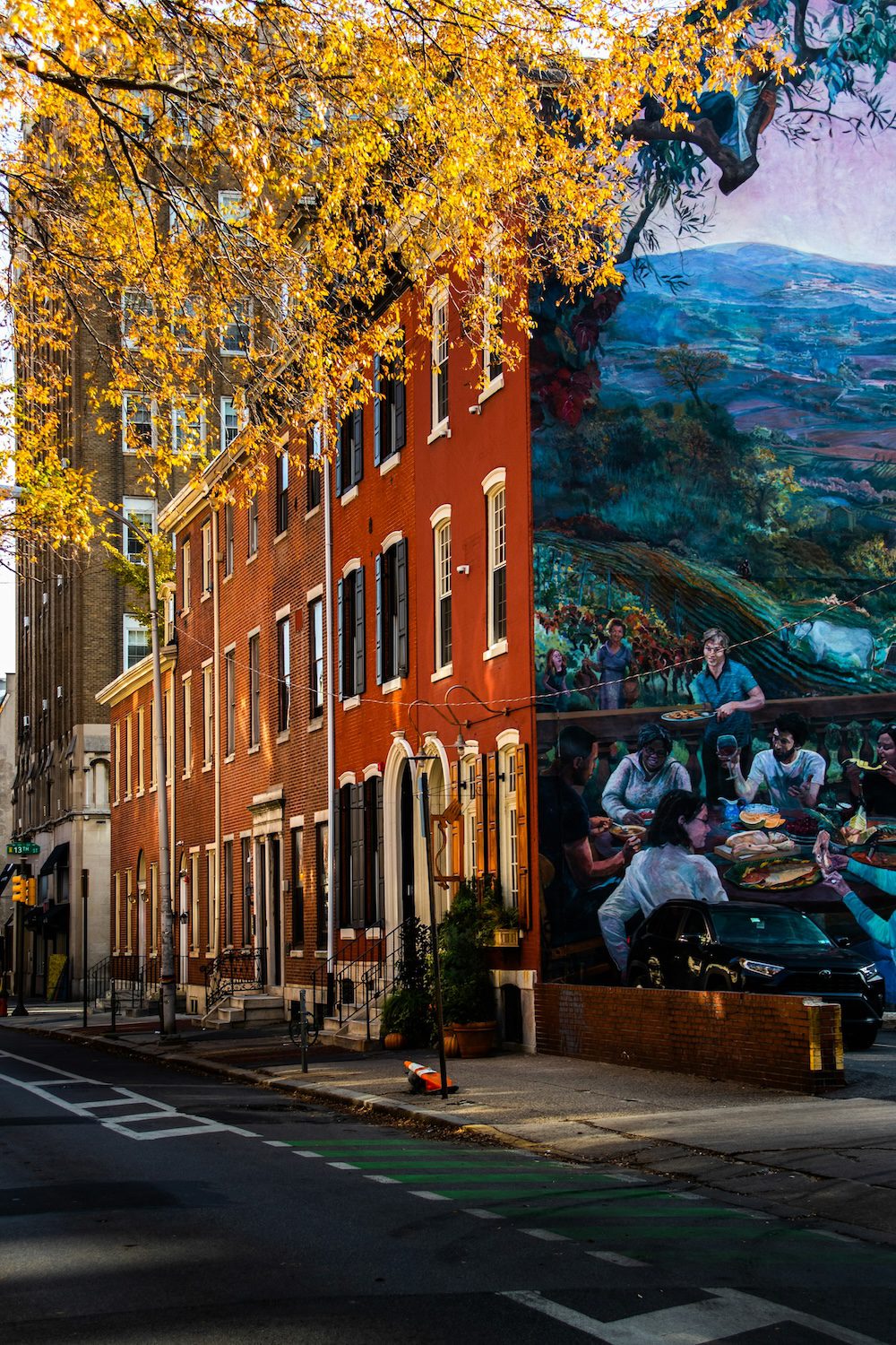 Large mural on the side of a brick building, likely a rowhome, in Philadelphia in fall, with visible yellow leaves on a tree in the foreground. The mural shows a rural landscape with mountains in the background, and a people sharing an outdoor meal or potluck in the foreground.