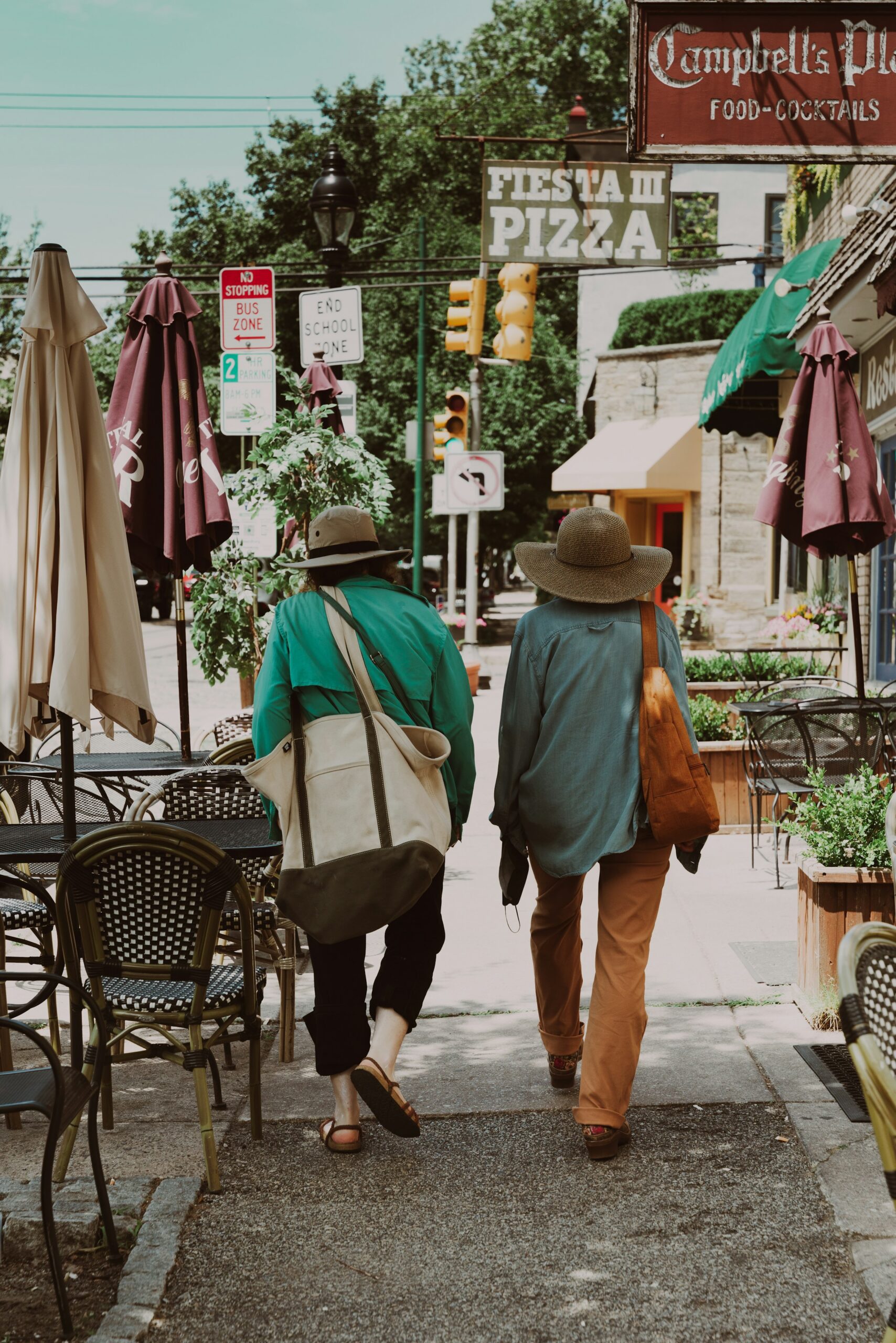 Two women walk on a sidewalk facing away from the photographer. It is sunny and they are dappled in shade from a non-visible tree. Both women wear shade hats and cary large tote bags. They are casually dressed, one is wearing sandals. They are passing restaurants with tables on the sidewalk. No diners at the tables, and the umbrellas are down. Ahead of them, signs on buildings read "Campbell's Place Food - Cocktails" and "Fiesta III Pizza." Several street signs are also visible.